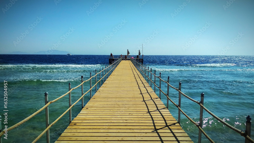 Obraz premium Long wooden pier leading into the clear turquoise sea under a bright blue sky. Idyllic vacation setting with a scenic view of the water and distant horizon.