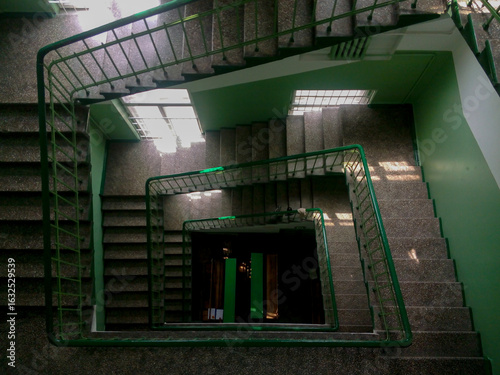 Top-down view of a spiral staircase with green railings inside a residential or office building. Geometric symmetry and architectural details.