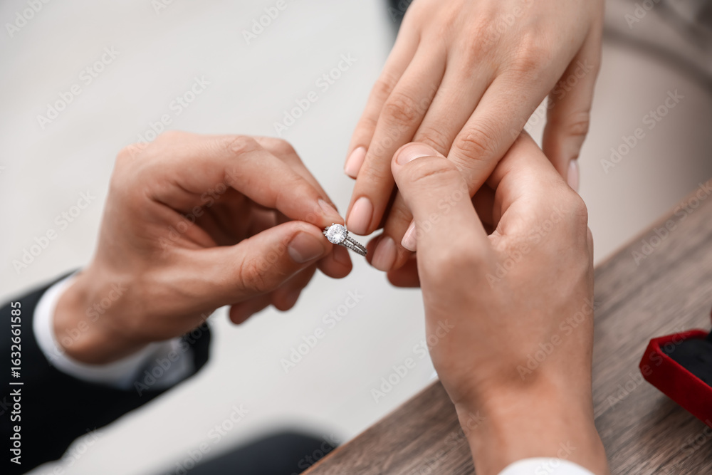 Fototapeta premium Making proposal. Man putting engagement ring on his girlfriend's finger indoors, closeup