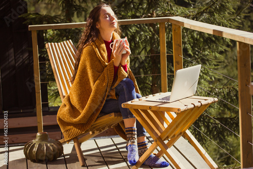 Relaxing woman on a wooden balcony enjoying coffee and working on laptop in the forest setting. A calm girl enjoys tea drink and sunlight on a wooden terrace, wrapped in a cozy blanket enjoying warmth