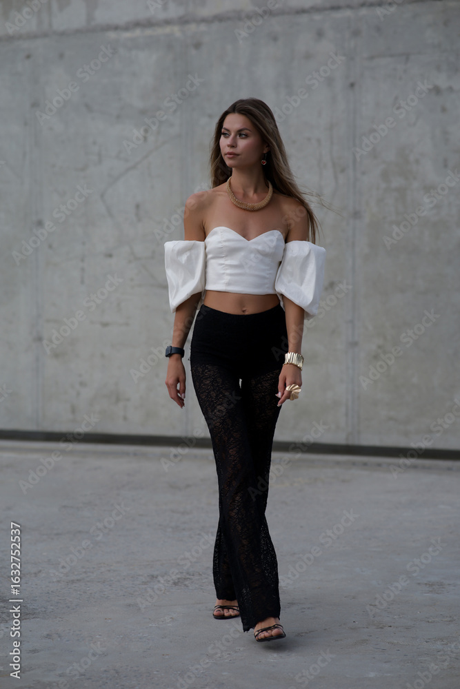 Fototapeta premium Stylish young woman in fashionable clothes posing confidently outdoors. Fashion model look with urban background, long hair, black shoes, and modern accessories.
