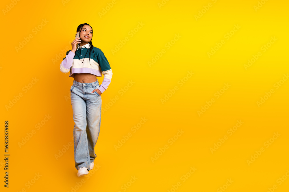 Fototapeta premium Cheerful young woman with braided hair talking on phone and posing confidently against a vibrant yellow background