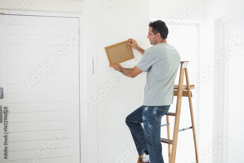 Wooden picture frame being leveled on white hallway wall using stepladder near shuttered window