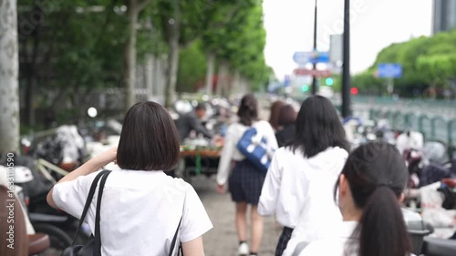 Slow-motion. Japanese high school girls in uniform buy and eat skewers cheerfully from a vendor, surrounded by trees and buildings blending in harmony. Spring. Shanghai's Pudong New District. China.