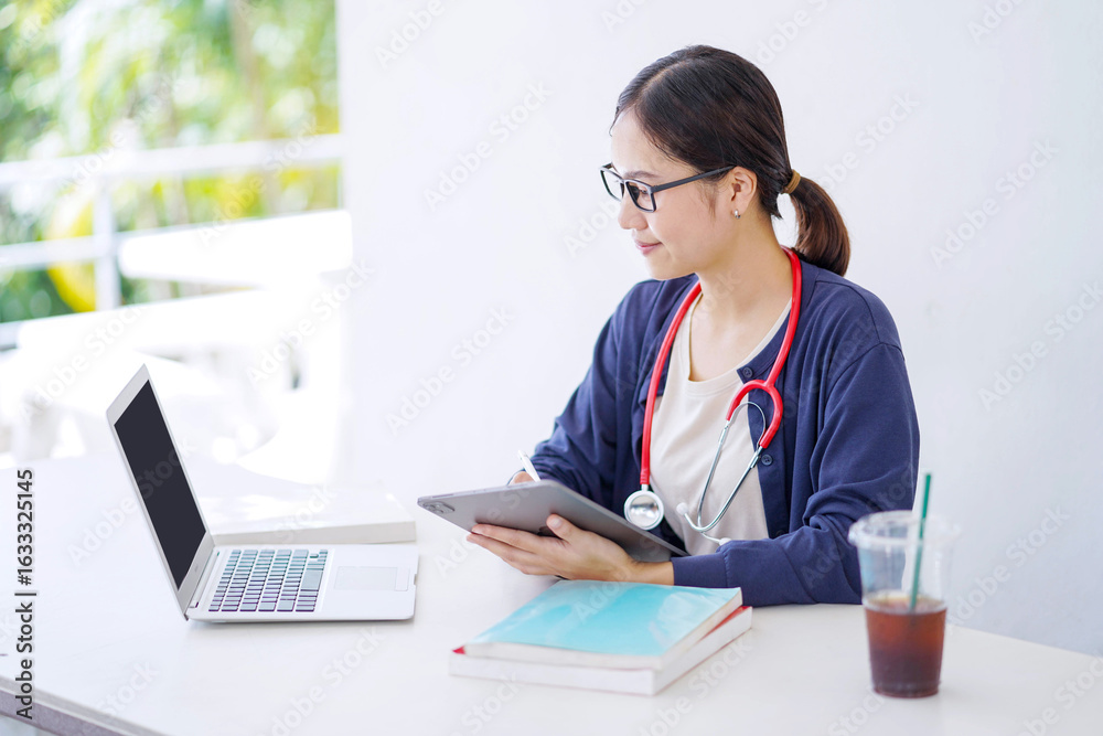 Asian young female medical student in casual cloth looking at laptop and make a short note on tablet in clinic building . Medical doctor concept. Asian young female medical student in casual cloth looking at laptop and make a short note on tablet in clinic building . Medical doctor concept.