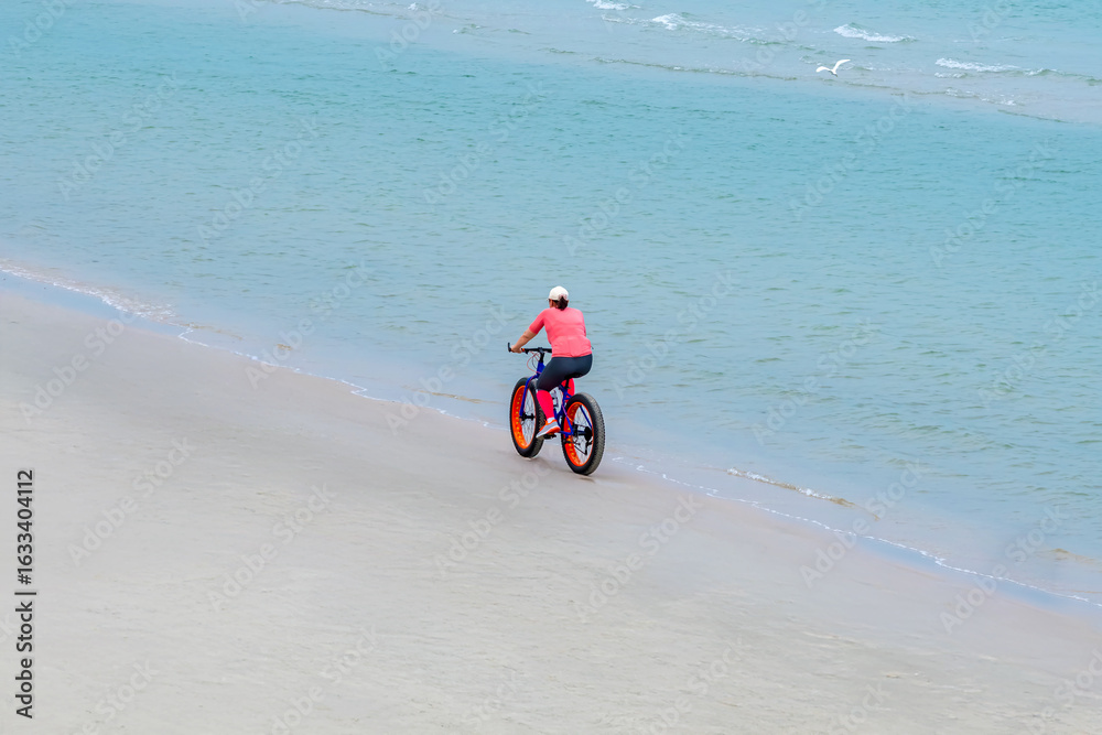 Obraz premium Carefree woman in sports fitness clothes riding Fat bike also called fatbike or fat-tire bike in summer driving on beach. Active healthy lifestyle. Enjoy and fun with bicycle on sand at coastal.