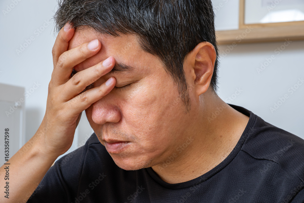 Fototapeta premium Middle-aged Asian man clutching his head, seated after long hours of work, experiencing headache and dizziness—ideal for medical ads on fatigue relief, neurology, wellness care.