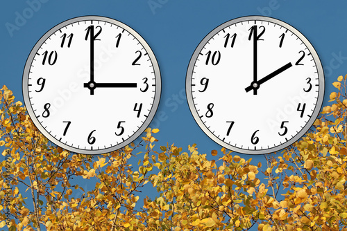 wo clocks, one showing three o’clock and the other showing two o’clock, against the sky and autumn yellow trees. A symbol of the time change from summer time to winter time, moving the hands