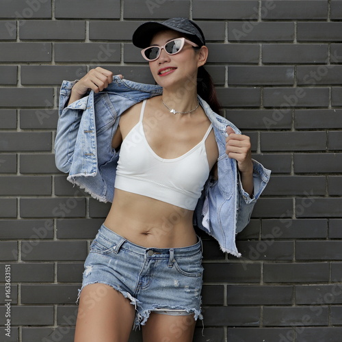 Effortlessly cool pretty Asian female embodies modern urban style in a sleek white crop top and denim shorts, with a confident stance against a textured wall