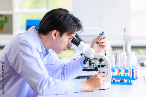 Asian man scientist medical development researcher chemistry examining samples under microscope pharmaceutical focus in lab coat working analyzing test tubes in modern laboratory equipment workspace.