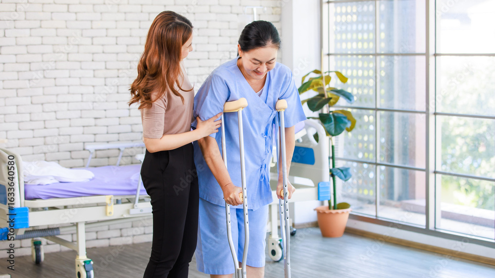 Fototapeta premium Asian young woman teaching using crutches walks beside senior female patient sitting on hospital bed medical recovery room caregiver specialist wellness healthcare treatment rehabilitation support.