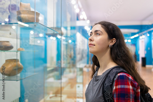 Portrait of woman in museum examining ancient clay vessels through glass, representing learning, heritage, and the importance of museums for public education