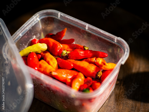 Close-up of assorted red and yellow chili peppers inside a transparent plastic container, capturing the freshness, heat, and vivid color under natural sunlight on a rustic surface.