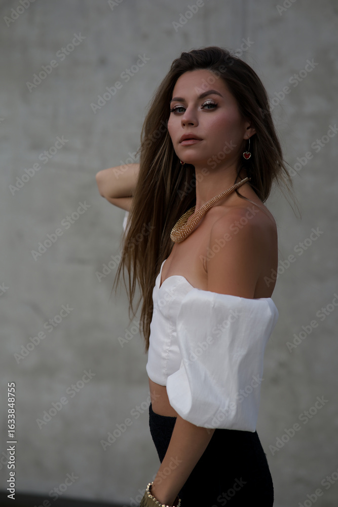 Naklejka premium Stylish young woman in fashionable clothes posing confidently outdoors. Fashion model look with urban background, long hair, black shoes, and modern accessories.