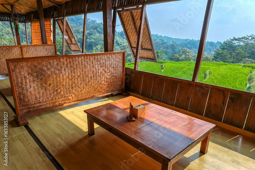 A rustic wooden interior with a table and woven bamboo partitions, overlooking a lush green rice paddy field and a dense forest landscape.