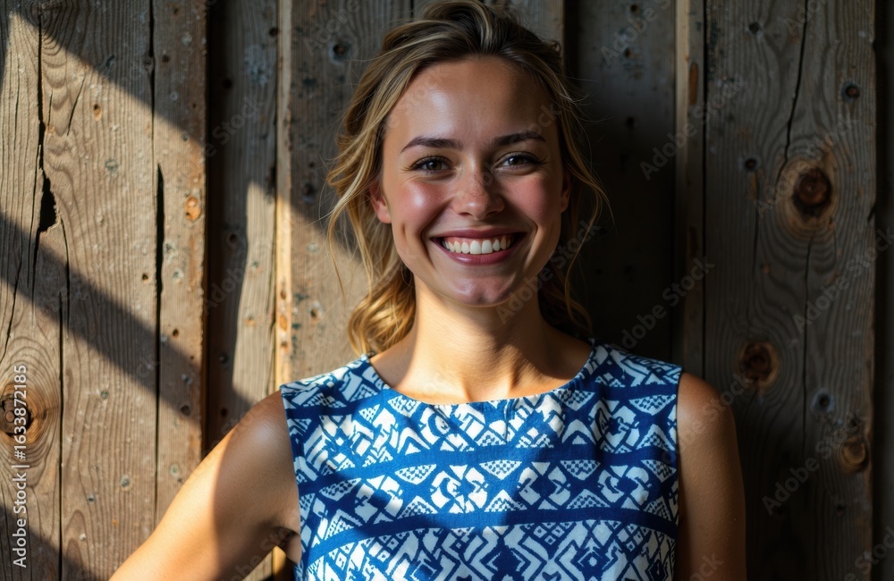 Fototapeta premium A smiling woman with blonde hair wearing a blue patterned dress standing outdoors in front of a wooden fence