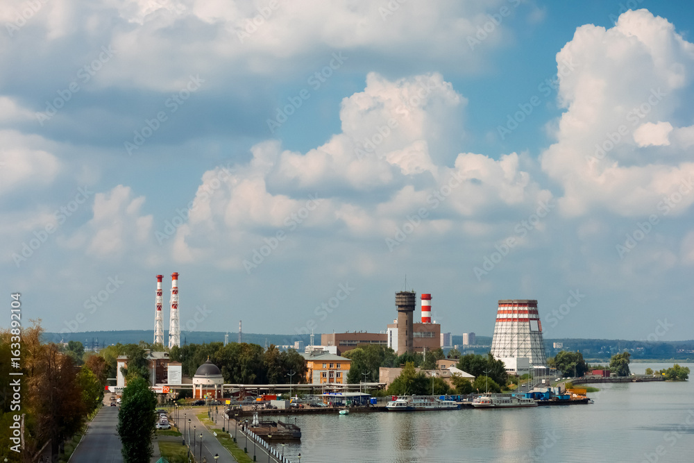 Naklejka premium On the river bank there is a thermal power plant with tall chimneys, surrounded by greenery. The sky is covered with fluffy clouds, creating a calm and picturesque view.
