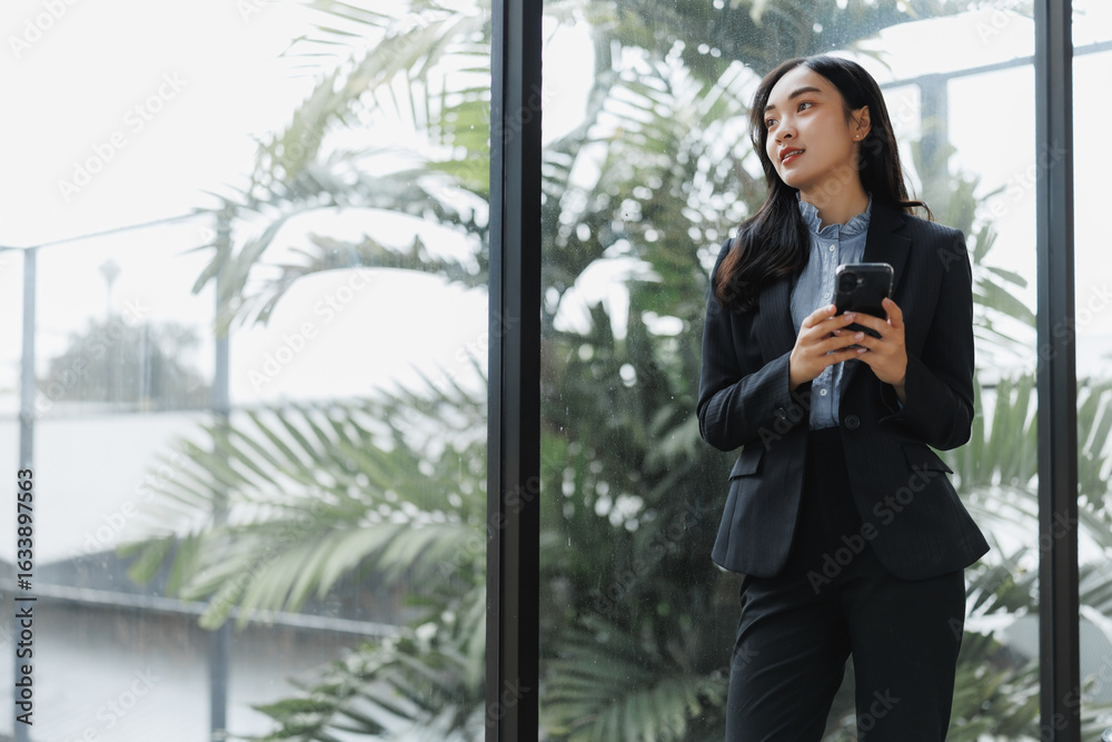Fototapeta premium Businesswoman holding smartphone and looking out large window in modern office