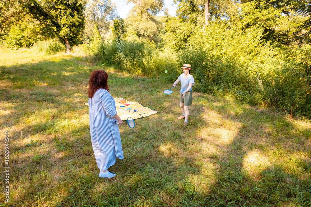 Fototapeta premium Caucasian woman and her daughter enjoy badminton in a green park with trees. Real people, outdoor game, bonding and holiday mood.