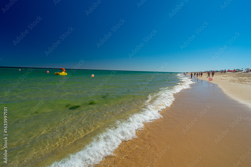 Obraz premium Relaxed summer holiday atmosphere with crystal clear sea waters and a yellow leisure boat with a red slide floating in the calm waters of Barril beach - Santa Luzia - Tavira - Algarve - Portugal