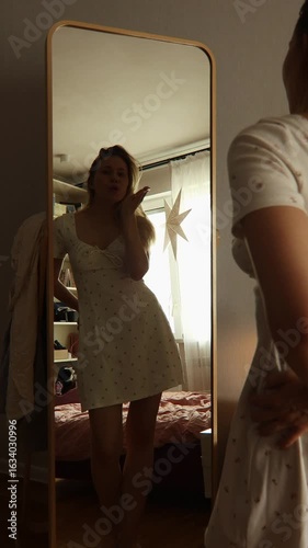 A young woman stands in front of a tall mirror, wearing a white dress. She showcases her style while playfully tossing her hair, capturing a moment of self-expression and confidence.