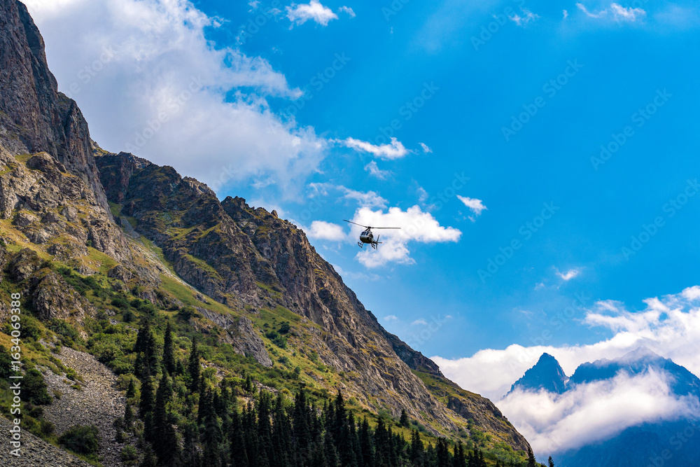 Fototapeta premium A light rescue helicopter is flying next to a mountainside against a blue sky with clouds and mountain peaks.