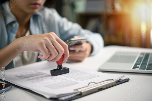 Fotografia Hand of a professional woman stamping official papers on a clipboard with a laptop and phone on her desk