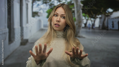 Blonde woman holds both hands up in a defensive gesture on a sunlit urban street; caution hesitation uncertainty restraint.