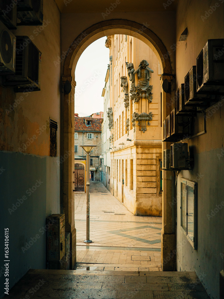 Fototapeta premium Historic stone passage in Republic Square, Split, Croatia, frames a narrow tiled street with ornate facades, leading to Neretvanska ulica under morning light.