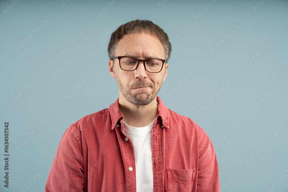 Fototapeta premium Upset thoughtful man sadly looks down biting lips with excitement frowning forehead wearing eyeglasses isolated background. Stressed nervous middle aged male worried thinking about troubles solutions.