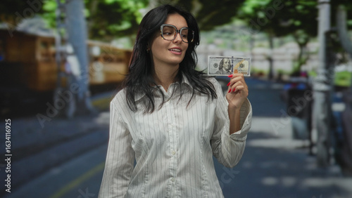Fotografija Woman wearing glasses holding hundred dollar bill on street paved sidewalk under green trees; wealth confidence