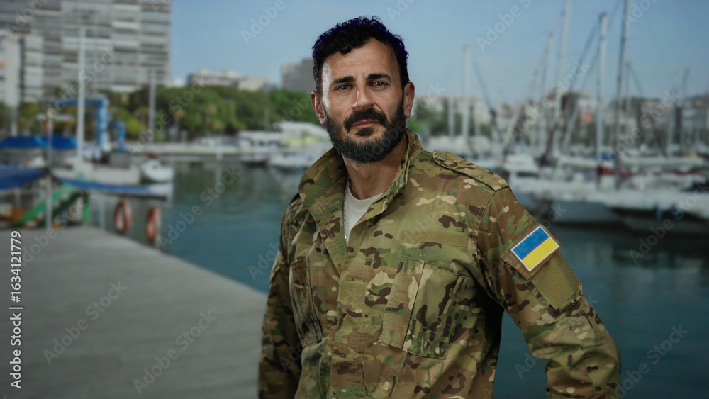 Fototapeta premium Hispanic man in military uniform with ukrainian flag patch stands near port with boats and ships, showcasing a maritime military scene with a calm sea backdrop.