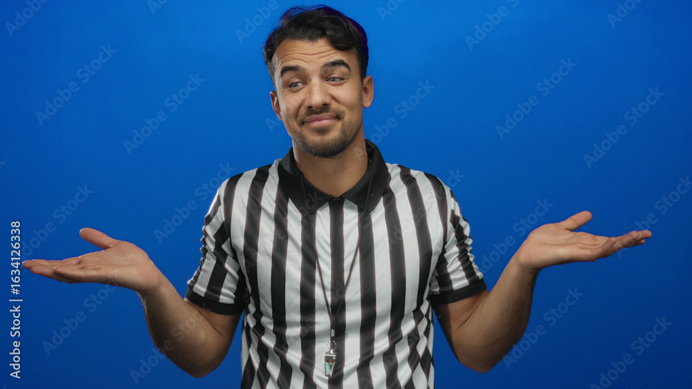 Fototapeta premium Young hispanic man referee in striped shirt gestures with uncertainty against an isolated blue background.