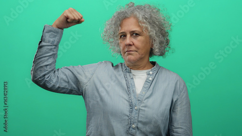 Canvas Print Elderly woman with grey hair does a strength gesture against an isolated green background wall, embodying empowerment and resilience