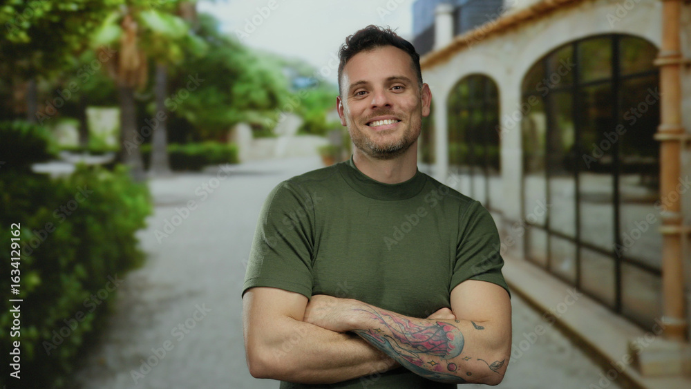 Fototapeta premium Smiling man with tattoos in a green shirt stands with crossed arms on a sunny urban street lined with trees and buildings creating a relaxed and confident atmosphere