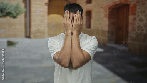 Man covers face with hands in a narrow cobbled old town street while standing under brick arches; embarrassment shame discomfort.