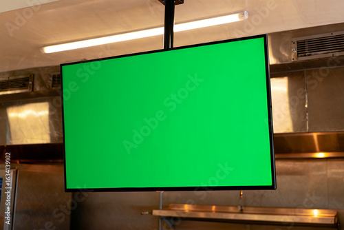 A ceiling-mounted television with a blank green screen for mockups hangs in a professional kitchen.
