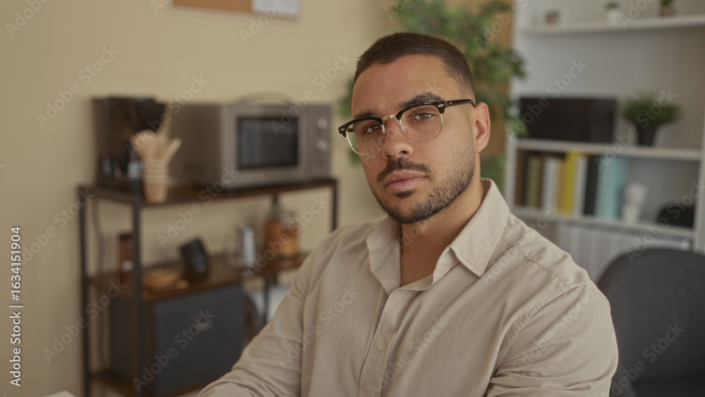 Fototapeta premium Young hispanic man wearing glasses glances to the side at a coffee station on a shelf in an office workspace; contemplation.