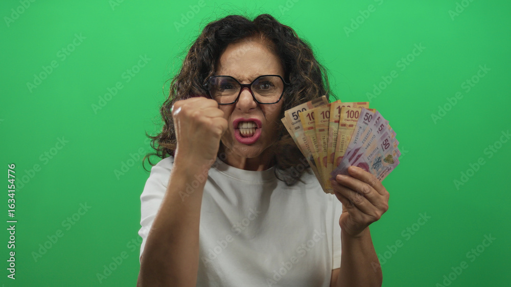 © Krakenimages.com - Middle aged woman wearing glasses holds mexican peso banknotes and shakes fist in studio; anger frustration financial stress.