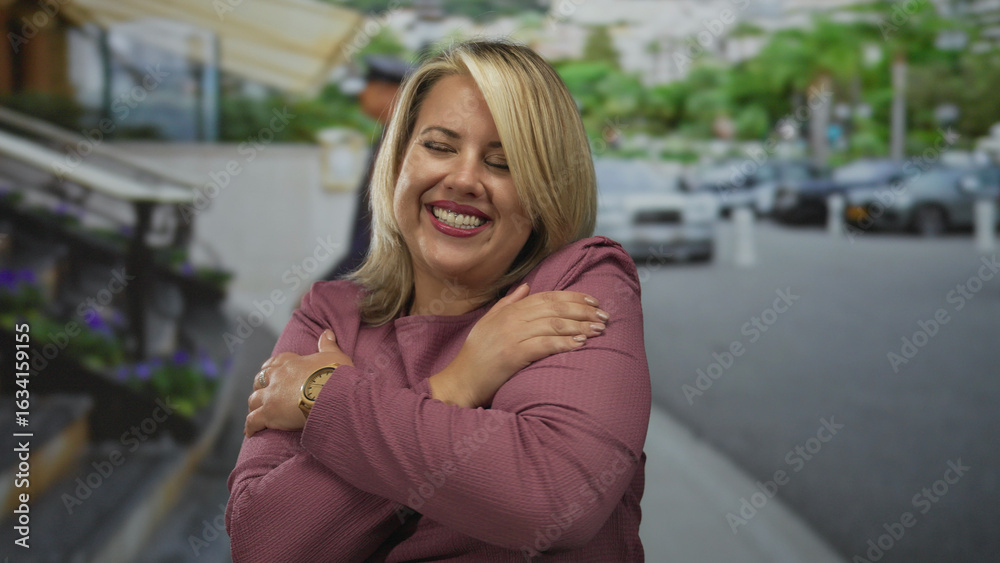 Fototapeta premium Blonde woman hugging herself on a busy outdoor city street while brightly smiling at the camera; self love.
