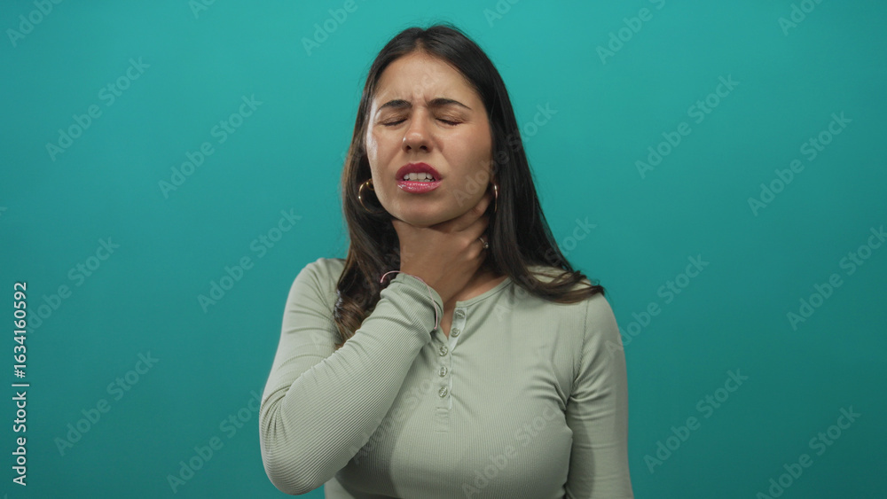 Fototapeta premium Young hispanic woman holds throat with both hands for a sore throat in a green studio; health concern discomfort.