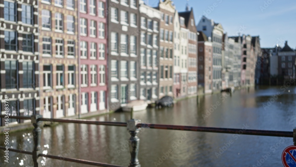 Naklejka premium Blurred view of amsterdam canals with defocused buildings and bokeh effect under a clear sky, creating a serene and picturesque dutch urban scene.