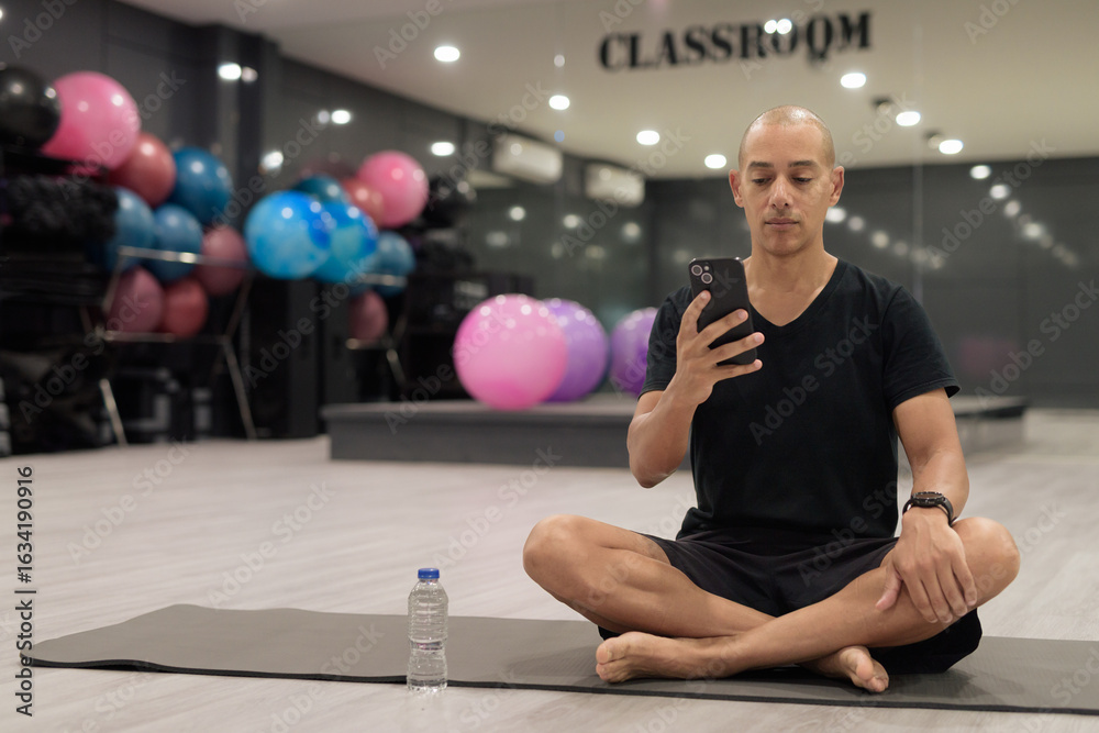 Fototapeta premium Hispanic bald man athlete in gym classroom doing yoga meditation while sitting and using phone