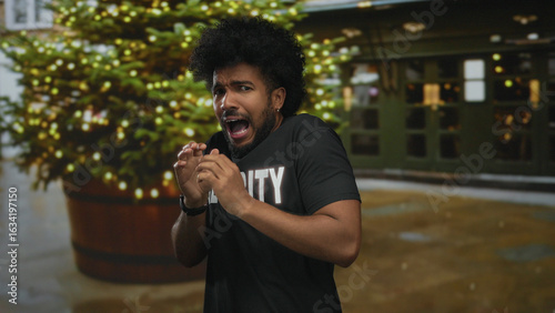 African american man in security uniform looks fearful on city street with blurred festive lights behind, conveying a tense outdoor scene.