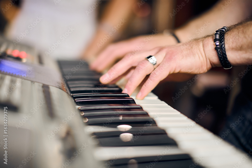 Obraz premium Close-up of adult male hands playing keyboard in vibrant music band rehearsal. Soft focus on background conveys dynamic atmosphere. Modern setting, wristband detail