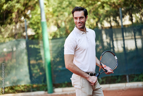 A cheerful man in a light-colored polo shirt and beige shorts stands ready on a tennis court. He holds a tennis racket and looks joyful under the warm sun