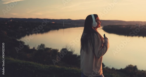 Young woman wearing headphones listening to music and dancing by the river at sunset during autumn