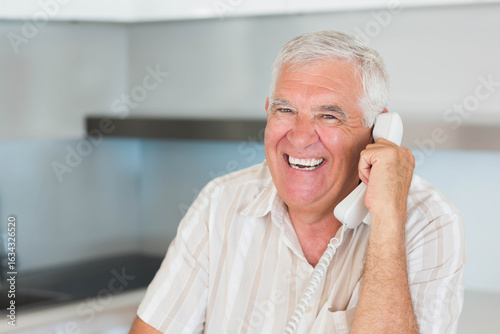 Senior man talking on corded telephone in modern kitchen with countertop and cabinets, copy space