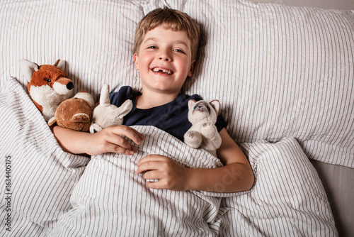 Happy child waking up in bed with big smile and raised arm, surrounded by stuffed animals. Joyful morning energy, childhood happiness and cozy home atmosphere. Fun before a sleeping. Kid smile in bed.