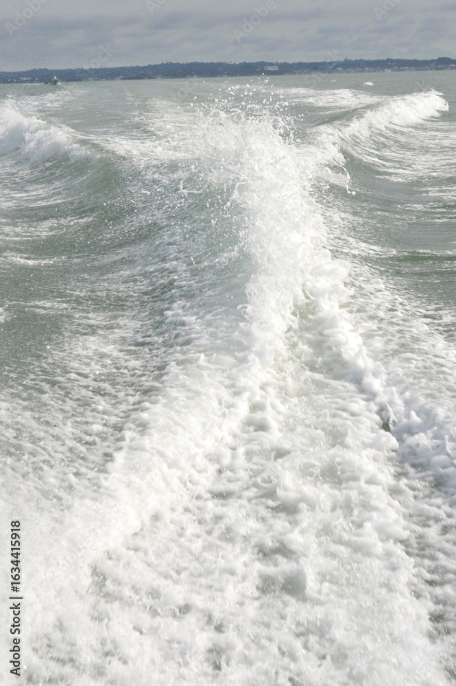 Fototapeta premium Dynamic ocean waves crash creating white foam trails behind a moving boat on a cloudy day